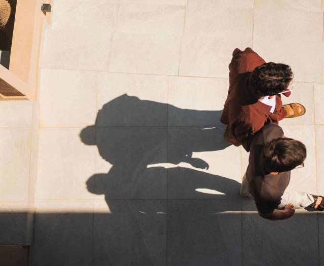 Two guests walking across a sunlit terrace at Paralos Venus Suites, casting long afternoon shadows.